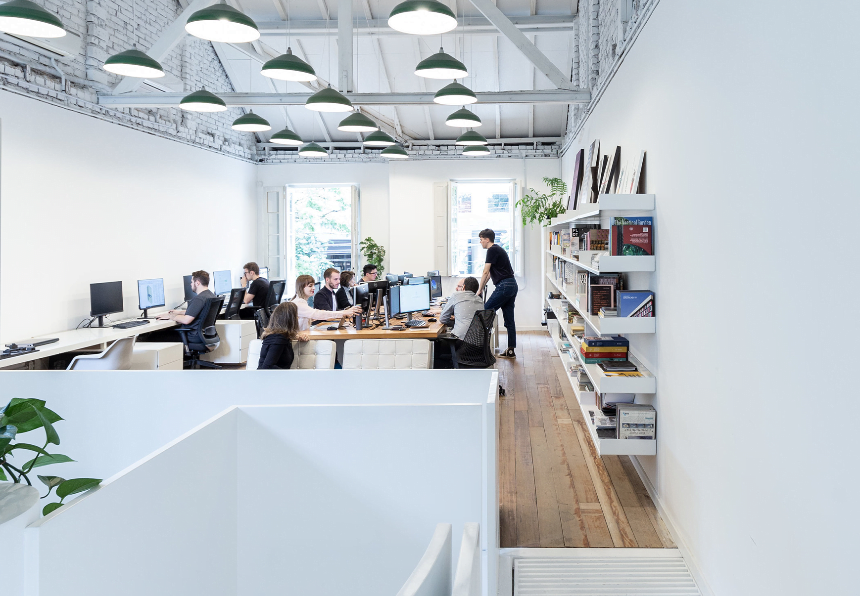 Uma equipe de pessoas trabalhando em computadores em um escritório moderno com paredes brancas, teto alto e luzes verdes suspensas. Estantes com livros e plantas são visíveis à direita.